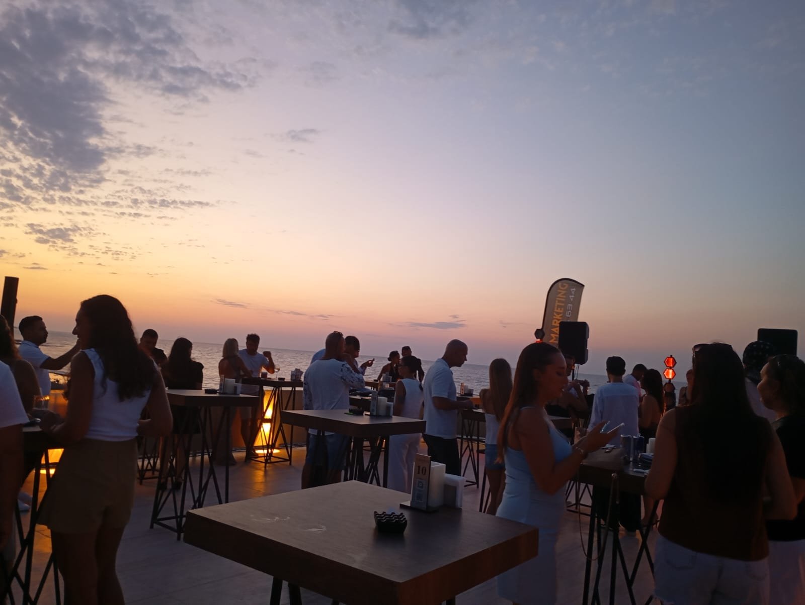 Sundowner-Party an der Beachbar bei Sonnenuntergang, Menschen an Stehtischen mit Blick aufs Meer