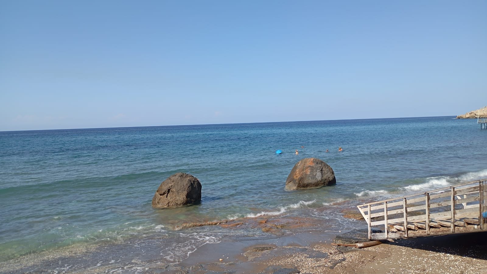 Strand auf Nordzypern: zwei große Felsen im flachen türkisfarbenen Meer, rechts ein Holzsteg, im Hintergrund Menschen im Wasser.