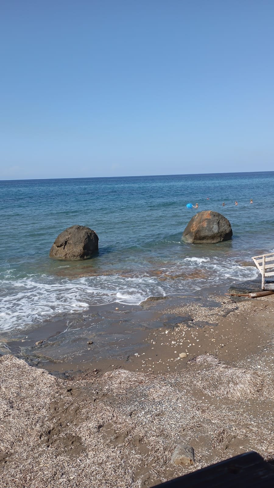 Näherer Blick auf die beiden Felsen im Meer, davor Sandstrand mit angespültem Seegras; im Wasser Menschen und ein blaues Etwas.